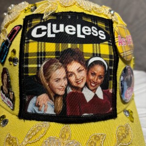 May include: Yellow baseball cap with a Clueless movie patch, featuring a plaid background and the movie title. The cap is embellished with pearls, rhinestones, and floral lace. Additional patches and buttons are also attached.