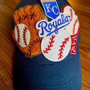 May include: A blue baseball cap with various embroidered patches, including a baseball glove, baseballs, and a patch with the text "KC Royals" in blue and white.