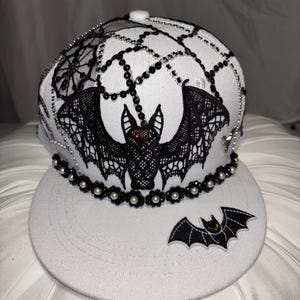 May include: A white baseball cap featuring a large black lace bat appliqué with orange eyes, surrounded by black and silver beaded spiderweb patterns. The brim is adorned with a smaller black bat patch and a row of black flower and white pearl beads.