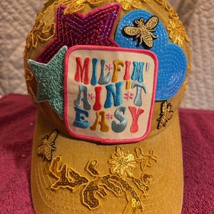 May include: Mustard yellow baseball cap with embroidered embellishments. Features a patch with the text "MILFIN' AIN'T EASY", along with star, bee, and floral designs. The brim and crown are adorned with sequins and gold embroidery.