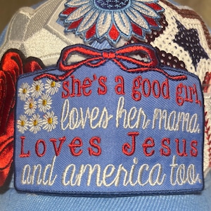 May include: A light blue baseball cap featuring a rectangular patch with the embroidered text 'she's a good girl loves her mama. Loves Jesus and america too.' surrounded by red, white, and blue floral and star-shaped embroidered patches.
