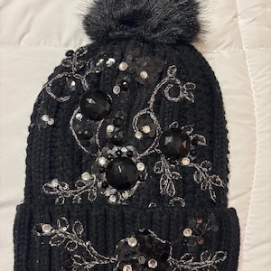 May include: Black knit beanie with a faux fur pom-pom on top. The hat is embellished with black sequins, silver beads, and large black faceted stones arranged in floral and vine patterns. A stylish winter accessory.