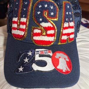 May include: A navy blue baseball cap featuring the letters USA in a textured American flag pattern on the front panels. The brim displays the number 50 with a Liberty Bell graphic, white stars, and a flag motif. The top is adorned with black sequins.