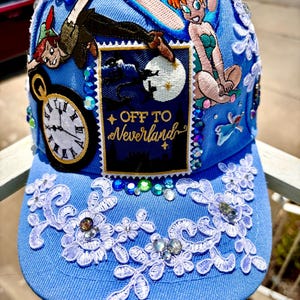 May include: A light blue baseball cap featuring Peter Pan and Tinkerbell designs, with the text "OFF TO Neverland". The cap is adorned with white lace, rhinestones, and a clock graphic. A whimsical accessory.