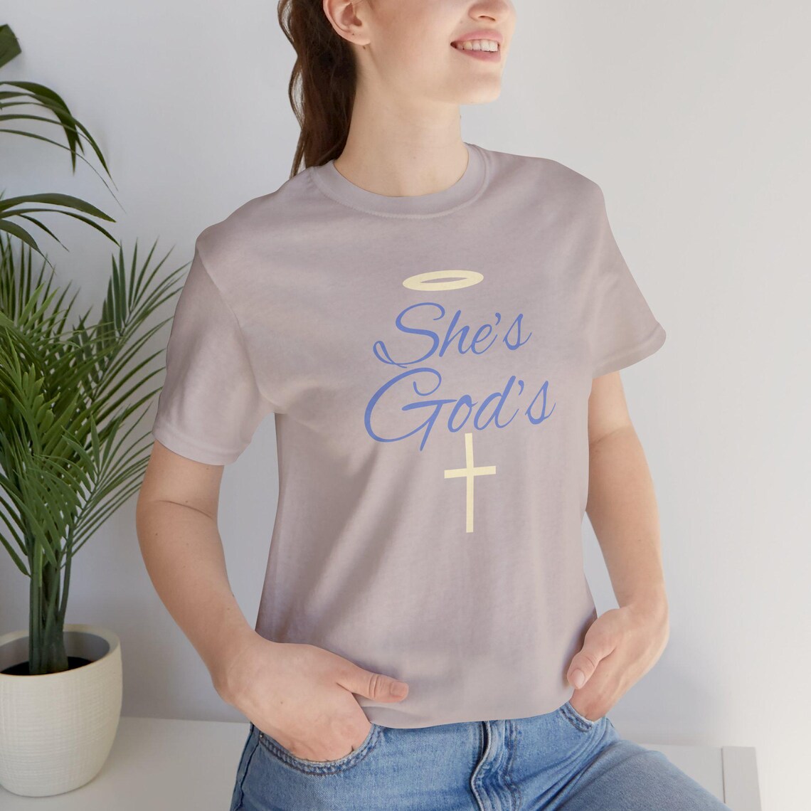 May include: A heather gray t-shirt with the text 'She's God's' in blue lettering with a yellow halo above the text and a yellow cross below the text.