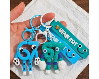 May include: Two silicone keychains featuring the number 67 as a cartoon character with large eyes and white gloves, attached to blue and teal rectangular tags with the text BRAIN ROT and a simple smiling face graphic, connected by metal rings.