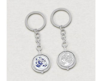 May include: Two silver keychains with round pendants. One pendant features a blue and white floral design, while the other has a silver floral design. Each keychain has a chain and a circular key ring.