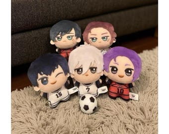 May include: Five plush dolls with anime-style features, including large eyes and colorful hair. The dolls are wearing soccer-themed outfits with numbers. A small soccer ball sits in front of one doll. The dolls are on a light brown rug.