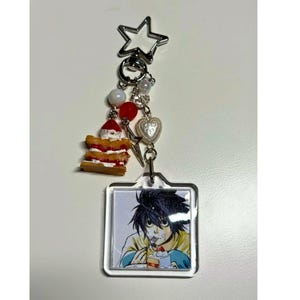 May include: A silver keychain with a star-shaped top, featuring a square charm with an anime character eating cake. The keychain includes a small cake charm, red and white beads, and a heart charm.