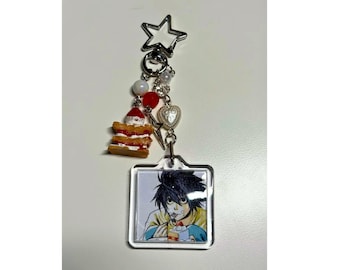 May include: A silver keychain with a star-shaped top, featuring a square charm with an anime character eating cake. The keychain includes a small cake charm, red and white beads, and a heart charm.