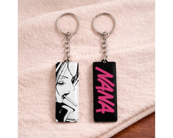 May include: Two keychains with silver key rings and chains. One keychain has a black and white illustration of a woman smoking. The other keychain has the word "NANA" in pink on a black background.