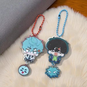 May include: Two anime-style keychains. One keychain has a red chain and features a character with blue hair and a flower. The other has a blue chain and a character with black hair. Both have a charm at the bottom.