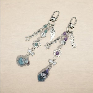 May include: Two keychains with silver-tone hardware and decorative charms. Each keychain features a dangling design with beads, stars, flowers, and a character illustration. One keychain has a blue-haired character, and the other has a witch.