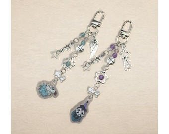 May include: Two keychains with silver-tone hardware and decorative charms. Each keychain features a dangling design with beads, stars, flowers, and a character illustration. One keychain has a blue-haired character, and the other has a witch.