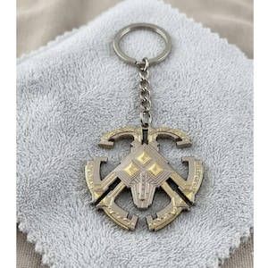 May include: A silver and gold-toned metal keyring with a decorative emblem. The emblem features geometric shapes and a central design with gold accents. The keyring is attached to a silver chain and ring.