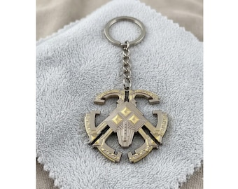 May include: A silver and gold-toned metal keychain with a decorative emblem. The emblem features geometric shapes and a central design with gold accents. The keychain is attached to a silver chain and ring.