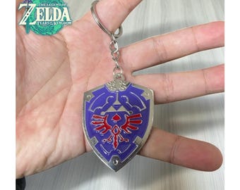 May include: A silver-colored keychain featuring a purple shield with red and white accents. The shield design includes a stylized emblem. The keychain is attached to a silver ring. The image also shows the text "The Legend of Zelda Tears of the Kingdom".