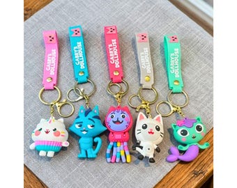 May include: Five silicone keychains featuring characters from Gabby's Dollhouse. Each keychain has a colorful strap with the text Gabby's Dollhouse, a gold-tone metal ring, and a unique character charm including a cat-cupcake, blue cat, pink octopus-cat, white cat, and green mermaid-cat.