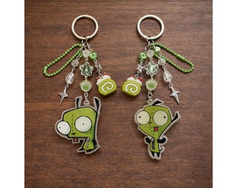 May include: Two keychains featuring green cartoon characters with large eyes and a small tongue. Each keychain has a silver ring, green beaded chain, and decorative charms including stars, flowers, and miniature cake rolls.