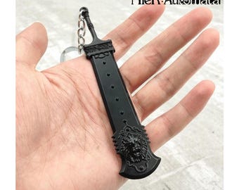 May include: A black sword-shaped keychain with a lion's head design on the hilt. The keychain has a chain and ring for attachment. The text "NieR:Automata" is visible in the upper right corner. The sword has a series of small circular indentations.