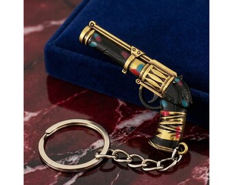 May include: A detailed keychain in the shape of a vintage pistol. The gun features gold, black, red, and blue accents. The keychain includes a silver-colored chain and a round key ring. The pistol is resting on a dark blue velvet surface.