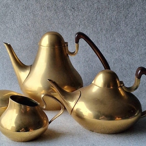 Könnte beinhalten: Ein goldfarbenes Teeset mit Teekanne, Milchkännchen und Zuckerdose. Die Teekanne hat einen braunen Griff und eine abgerundete, bauchige Form. Das Milchkännchen und die Zuckerdose sind ebenfalls abgerundet und haben eine glatte, polierte Oberfläche.