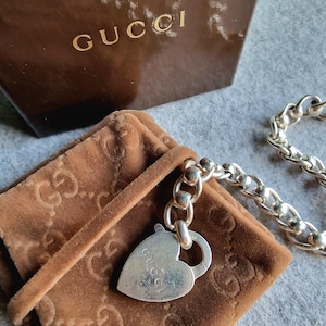 Könnte beinhalten: Eine silberne Kettenkette mit einem herzförmigen Anhänger, auf dem "Gucci" steht. Die Kette liegt auf einem braunen Samtbeutel mit dem Gucci-Logo.