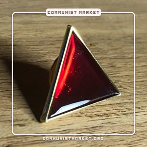 May include: A gold-colored triangular pin with a red glass center. The pin features a red, reflective, glass-like surface.