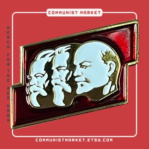 May include: A red, white, and gold enamel pin featuring three profile portraits of men. The pin is shaped like a rectangle with rounded corners and has the text "COMMUNIST MARKET" at the top and "COMMUNISTMARKET.ETSY.COM" at the bottom.