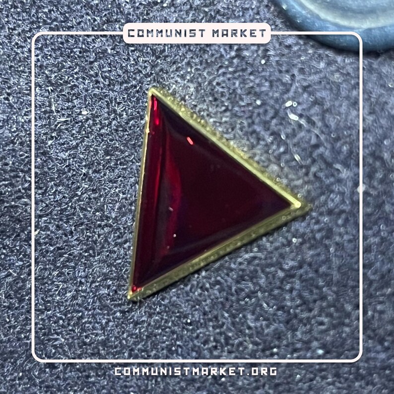 May include: A red triangle pin with a gold border. The pin is on a gray textured surface.