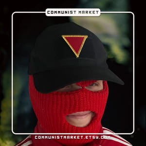 May include: A black baseball cap with a red triangle patch and gold trim. The cap is worn by a person wearing a red balaclava. The text "COMMUNIST MARKET.ETSY.COM" is visible at the bottom of the image.