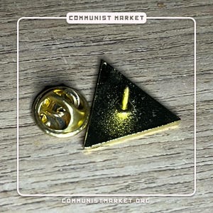 May include: A gold-colored triangle pin with a black background and a pin back. The pin is on a wooden surface.