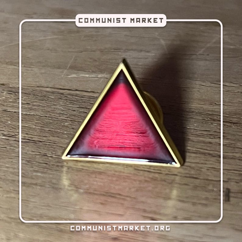 May include: A gold-colored triangle pin with a red and white design. The pin features a triangle shape with a red interior and a white border. The pin is on a wooden surface.