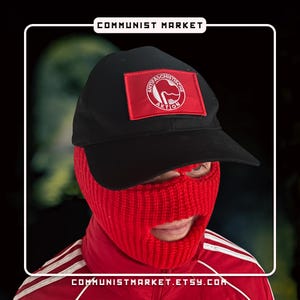 May include: A black baseball cap with a red patch featuring a white anti-fascist symbol and the text "Anti-Faschistische Aktion". The person is wearing a red balaclava.