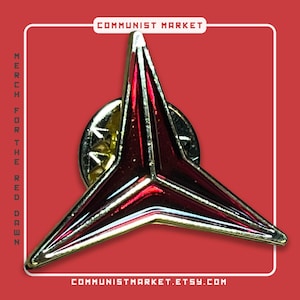 May include: A red and gold pin badge featuring a starburst design. The pin has a gold border and a red centre. The text "COMMUNIST MARKET" and "MERCH FOR THE RED DAWN" are visible. The Etsy shop URL is also visible.