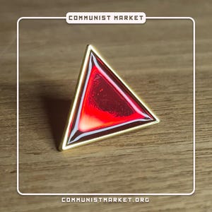 May include: A gold-colored triangle pin with a red, translucent center. The pin is on a wooden surface.