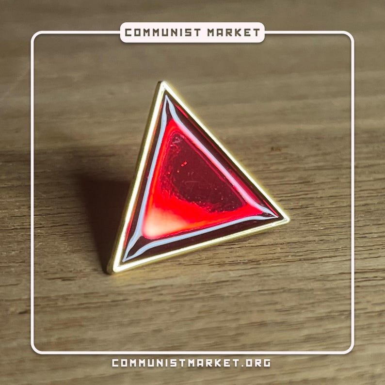 May include: A gold-colored triangle pin with a red, translucent center. The pin is on a wooden surface.