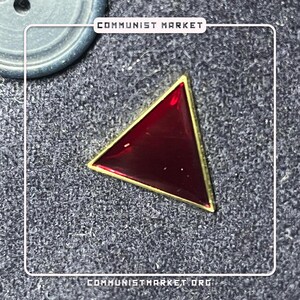 May include: A red triangle pin with a gold border. The pin is on a gray textured surface.