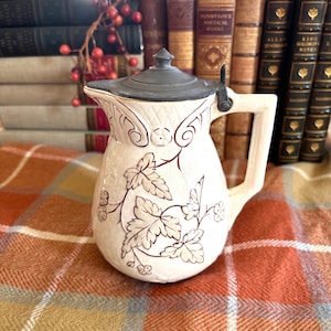 May include: Cream-coloured antique jug with a pewter lid and handle. The jug is decorated with a floral and leaf pattern in a dark reddish-brown colour. The jug is sitting on a plaid blanket.