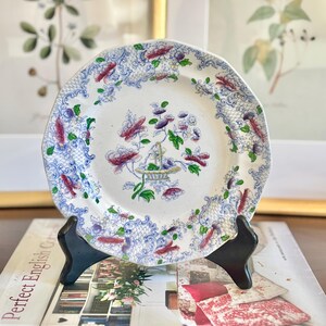 May include: A white porcelain plate with a blue and pink floral design. The plate has a scalloped edge and is decorated with a delicate floral pattern. The plate is on a black stand.