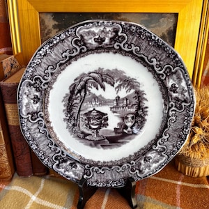 May include: A round ceramic plate featuring a black and white transferware pattern. The center depicts a landscape with a palm tree, a large urn, and buildings. The wide rim is decorated with an intricate scrollwork and floral border.