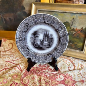 May include: A vintage decorative plate featuring a black and white landscape scene. The plate has a grey floral border and is displayed on a black stand. The plate's design is reminiscent of classic tableware.