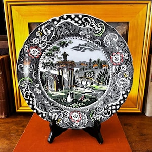 May include: A decorative plate with a black and white floral border and a central landscape scene. The plate features a scalloped edge and is displayed on a black stand. The landscape scene includes buildings, trees, and figures, with accents of red and green.