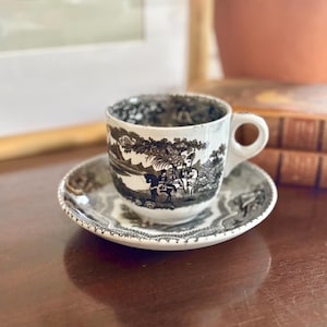 May include: A black and white teacup and saucer set. The cup and saucer feature a detailed pastoral scene. The cup has a small handle. The set is placed on a wooden surface.