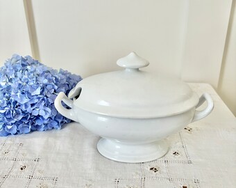 May include: White porcelain soup tureen with a lid and two handles. The tureen is sitting on a white tablecloth with an embroidered design.
