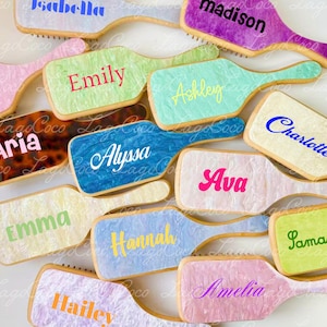 May include: A collection of personalized hairbrush-shaped cookies with names like "Isabella," "Madison," and "Sophia" in various colors. The cookies have a light-colored base with colorful text, and are arranged on a white surface.