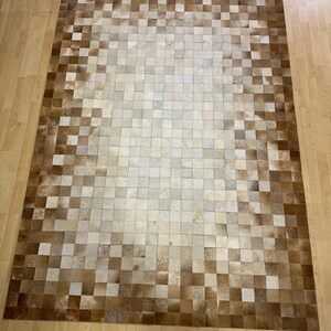 Könnte beinhalten: Ein brauner und weißer Patchwork-Rinderfellteppich mit quadratischem Muster. Der Teppich besteht aus einzelnen Stücken Rinderfell, die zusammengenäht wurden.