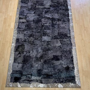 Pode incluir: Um tapete retangular com um design de patchwork em tons de cinza e preto. O tapete tem uma aparência texturizada e felpuda e é contornado por uma borda prateada com um padrão salpicado. O tapete está colocado sobre um chão de madeira clara.