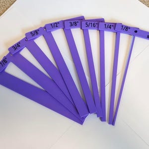 May include: A set of purple T-squares, each labeled with inch measurements from 1/8" to 1". The tools are arranged in a fan shape, with the text "HPS" visible on one. They are on a white surface.