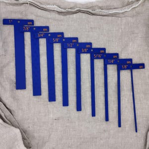 May include: A set of blue measuring tools with orange text indicating inch measurements from 1" to 1/8". Each tool has "HPS" printed on it. The tools are arranged in descending order on a neutral-colored fabric.